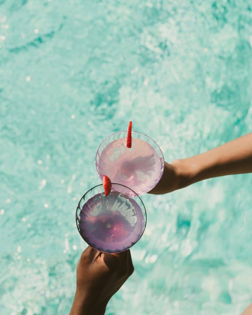 2 hands hold 2 big cocktail glasses in a clink over a summer pool, demonstrating large batch party cocktails for summer parties