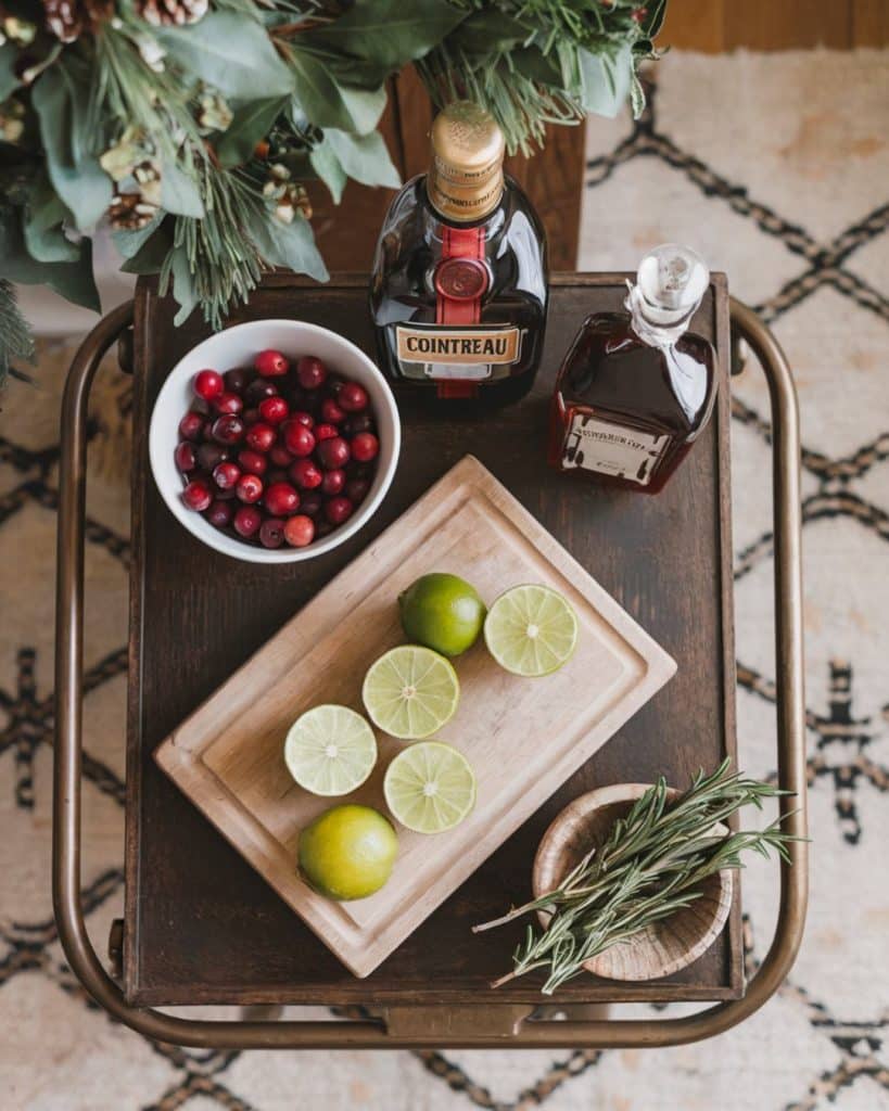 ingredients for a christmas margarita including cranberries, limes, triple sec, and tequila on a bar cart