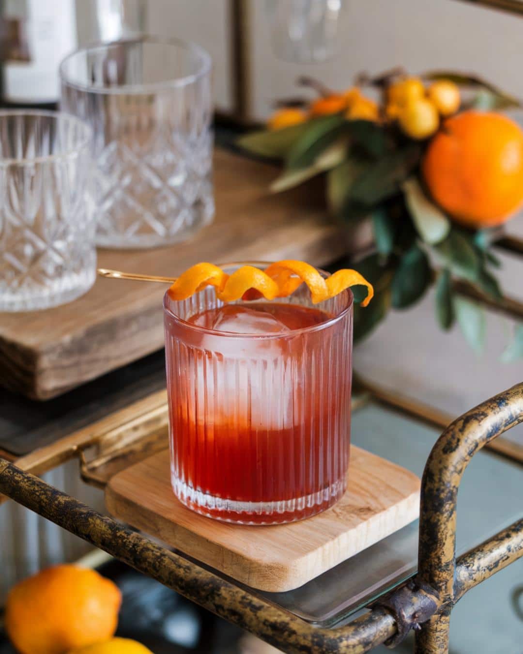 a negroni cocktail in a lowball glass with a large ice cube garnished with an orange twist sitting on a boho bar cart